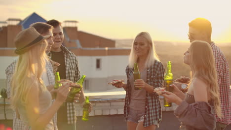 The-company-of-six-young-people-enjoy-the-time-on-the-roof.-They-celebrate-a-friends-birthday.-They-eat-hot-pizza-and-drink-beer-and-enjoy-smiles-in-the-summer-evening.-They-clink-beer-in-the-green-bottles-and-communicate-with-each-other.